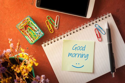 "Good morning" is written on opened notepad with pen, small sticky paper, smartphone, and paper clips on red cement background with morning scene