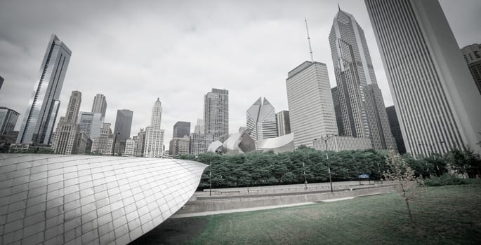 Skating Ribbon through Chicago's Millennium Park projects into frame below the city's highrise cityscape forms a towering background on an overcast grey day emphasised by desaturated image.