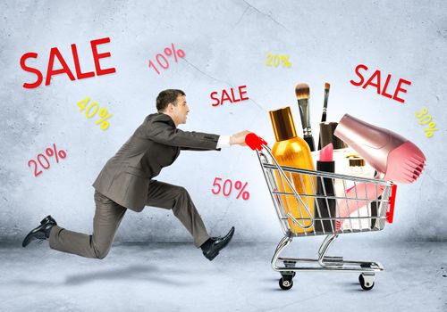 Businessman with cosmetics in shopping cart on grey background