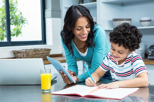 Kind mother helping her son doing homework in kitchen while using tablet and laptop