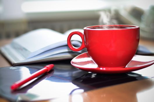 Red cup of hot coffee, notepad and pencil on the desktop in the office. 
Focus on the cup.