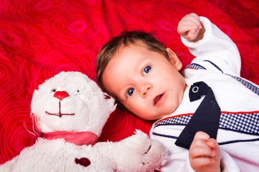 Portrait of newborn boy with blue eyes in her cozy red bed. With teddy bear. Lovely toy