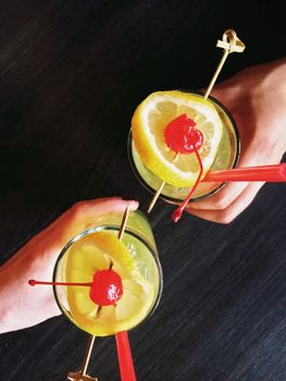 Hands of two people holding glasses with cocktail, decorated with lemon and cherry.