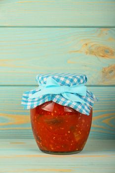 One glass jar of homemade pickled pepper, paprika and eggplant salad with checkered textile top decoration at blue painted vintage wooden surface