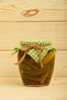 One glass jar of homemade pickled cucumbers with green checkered textile top decoration at beige painted vintage wooden surface