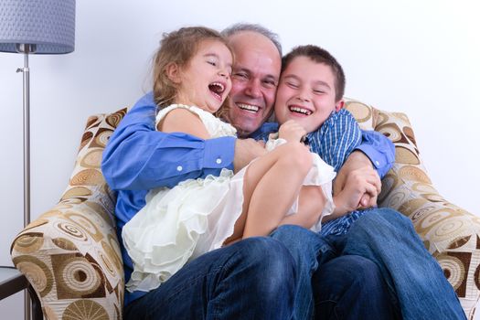 Middle-aged father with two laughing young kids, a girl and boy, on his lap sitting in an armchair enjoying a moment of fun and hilarity as a family