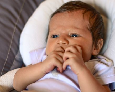 Thoughtful newborn boy looking left side with concentrated look and hand in the mouth,in his crib.