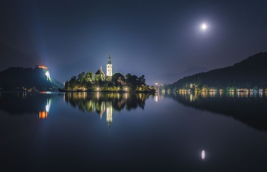 Illuminated Catholic Church on a Little Island and Bled Castle on Bled Lake in Slovenia at Night Reflected on Water Surface