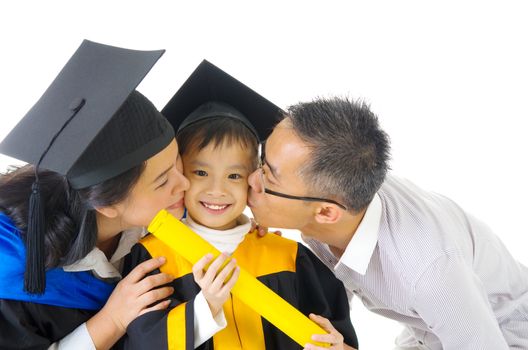 Asian kindergarten child in graduation gown and mortarboard kissed by her parent during graduation