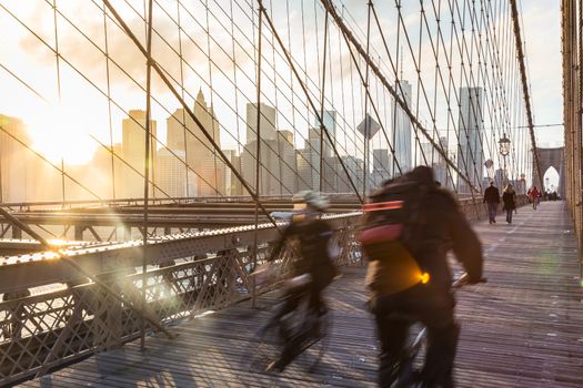 Cyclists cycle across Brooklyn bridge. New York City Manhattan downtown skyline in sunset with skyscrapers illuminated over East River panorama as seen from Brooklyn bridge.