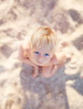 Little boy with blue eyes and white hair sitting on white sand and look up at the camera