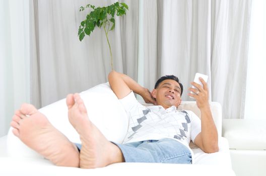 Smartphone mobile apps concept. Malay guy using smart phone. Handsome asian man relaxed and lying on sofa indoor.