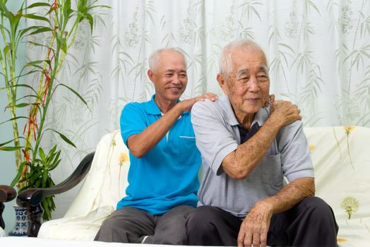 Asian old man shoulder pain, sitting on sofa , son massaging father shoulder. Chinese family, senior retiree indoors living lifestyle at home.
