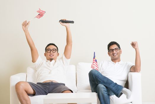 Young men watching live sport television program at home, waving Malaysia flag shouting and celebrate.