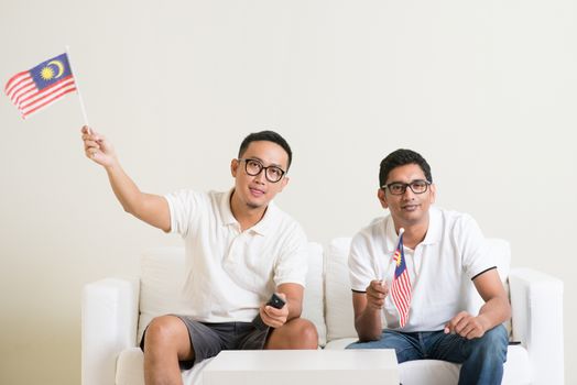 Young men watching live sport television program at home, waving Malaysia flag.
