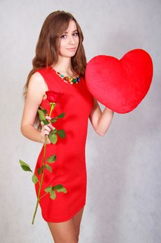 Portrait of a young attractive woman with a heart-shaped pillow and a rose over grey background