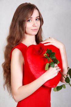 Portrait of a young attractive woman with a heart-shaped pillow and a rose over grey background