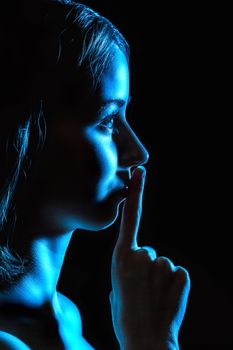 Contour of a beautiful young woman in blue light showing the silence sign over black background