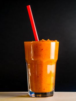 Simple still life with carrot smoothie in tall glass and red straw over black background, healthy life-style, dieting, eating imagery, modern look