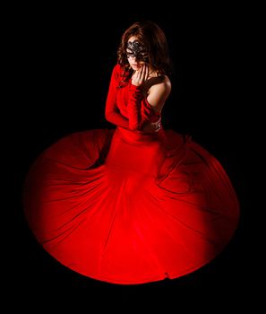 Beautiful young woman sitting in a long red evening dress over black background