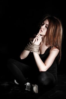 Beautiful young woman with tied arms sitting on the floor over black background