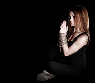 Beautiful young woman with tied arms sitting on the floor over black background