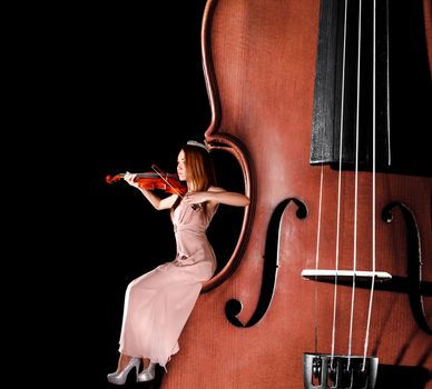 Tiny female violinist playing a violin sitting on the edge of the big one over black background