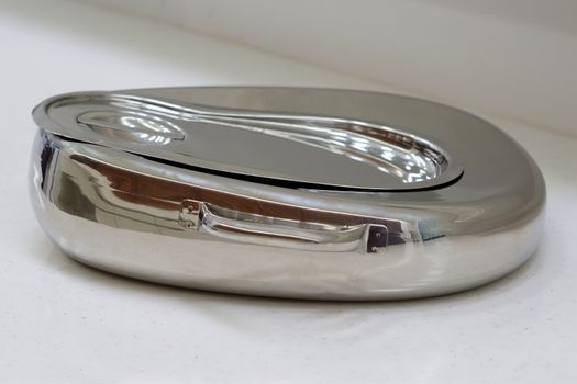  Stainless steel bedpan with cover placed on counter in hospital.                             