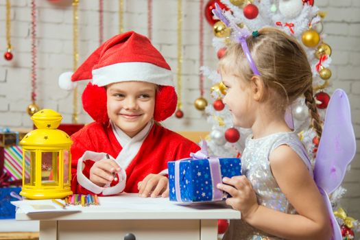 A girl dressed in a red suit Santa Claus draws pencils sitting at the table, next is a girl dressed as a fairy with the candlestick