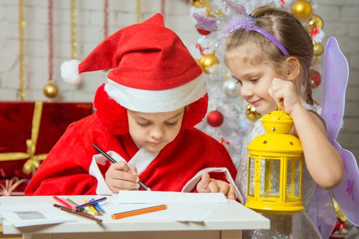 A girl dressed in a red suit Santa Claus draws pencils sitting at the table, next is a girl dressed as a fairy with the candlestick