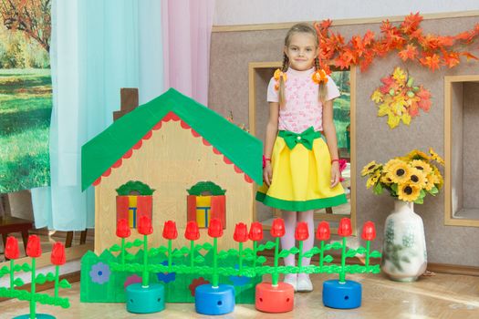 Six year old girl stands in the scenery of a country house and a flower bed at the matinee in kindergarten