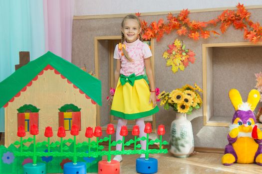 Six year old girl stands in the scenery of a country house and a flower bed at the matinee in kindergarten