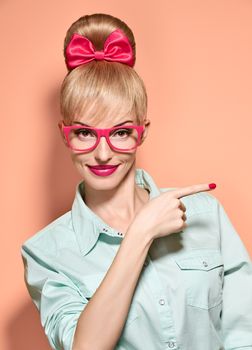 Beauty fashion nerdy woman, stylish glasses shows finger, idea. Attractive pretty funny blonde girl smiling. Confidence, success,Pinup hairstyle bow makeup.Unusual playful, expression.Vintage, on pink
