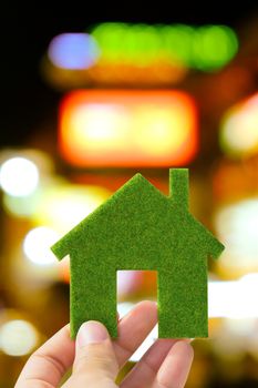 Hand holding small green eco house icon with defocused city night light background, green energy concept