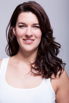 Smiling attractive woman with a lovely warm friendly smile and shoulder length curly brown hair looking at the camera over a grey background