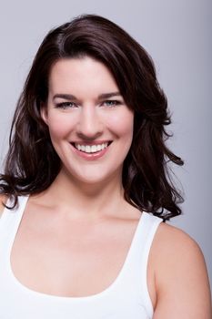 Smiling attractive woman with a lovely warm friendly smile and shoulder length curly brown hair looking at the camera over a grey background