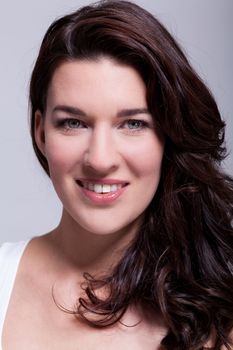 Smiling attractive woman with a lovely warm friendly smile and shoulder length curly brown hair looking at the camera over a grey background