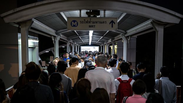 Bangkok, Thailand - January 18, 2016 : Crowd of people walking out from the BTS Mo Chit station to the road in the rush hour at night. They have to walk cross a footbridge along with lights and advertising board and the sign of the connected train above the head. Daily passengers of BTS skytrain is around 700,000.