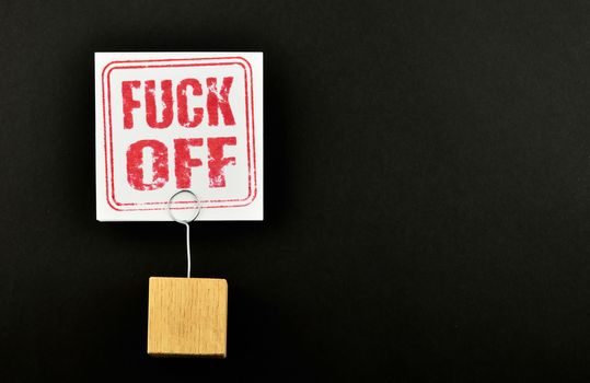 Fuck off, one white paper note with wooden holder isolated at black paper background for presentation