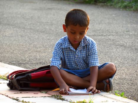 A little Indian boy sits down on a roadside footpath to study before his exam.                               