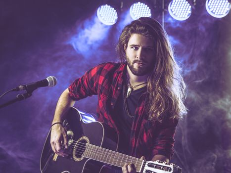 Photo of a young man with long hair and a beard playing an acoustic guitar on stage with lights and concert atmosphere.
