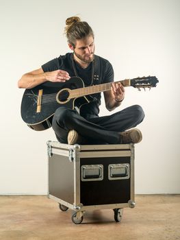 Photo of a young attractive man with long hair and beard sitting on a flight case and playing an acoustic guitar.