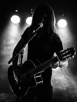 Photo of a backlit young man with long hair in silhouette playing an acoustic guitar on stage.
