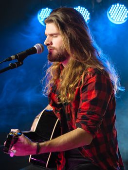 Photo of a young man with long hair and a beard singing and playing an acoustic guitar on stage with lights and concert atmosphere.
