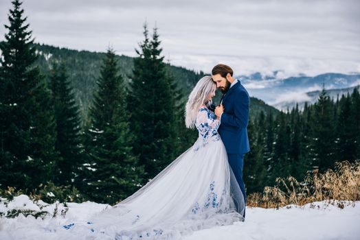 groom in a blue suit and bride in white, embroidered with blue pattern, dress on a background of green pine forests in the mountains of the Carpathians