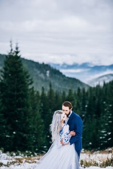 groom in a blue suit and bride in white, embroidered with blue pattern, dress on a background of green pine forests in the mountains of the Carpathians