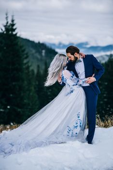 groom in a blue suit and bride in white, embroidered with blue pattern, dress on a background of green pine forests in the mountains of the Carpathians