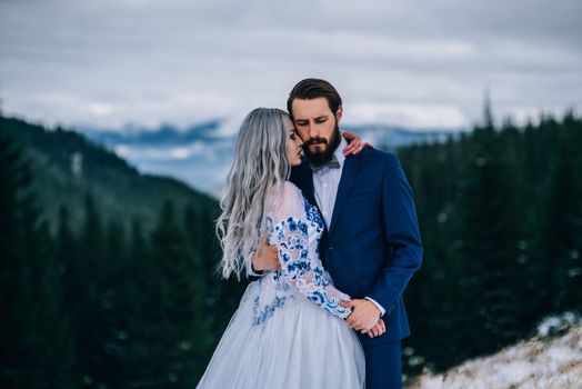 groom in a blue suit and bride in white, embroidered with blue pattern, dress on a background of green pine forests in the mountains of the Carpathians