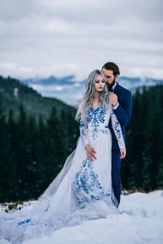 groom in a blue suit and bride in white, embroidered with blue pattern, dress on a background of green pine forests in the mountains of the Carpathians