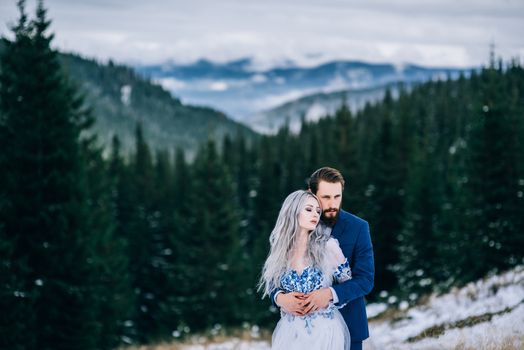 groom in a blue suit and bride in white, embroidered with blue pattern, dress on a background of green pine forests in the mountains of the Carpathians